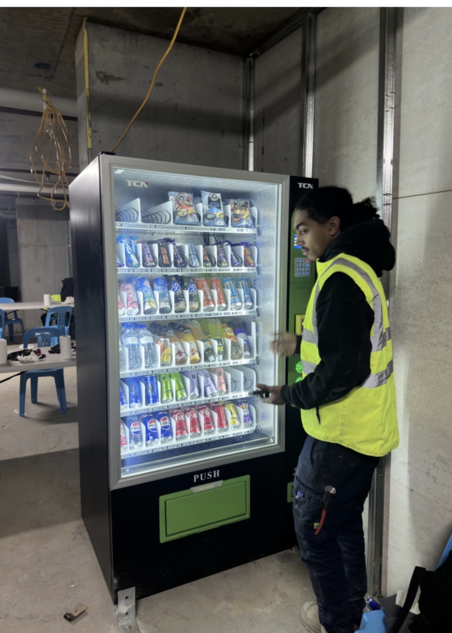 Worker beside a stocked Vendor Bros. vending machine at a job site